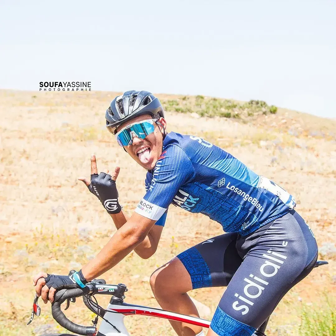 Sidi Ali cyclist smiling and making a hand gesture while riding through a dry landscape